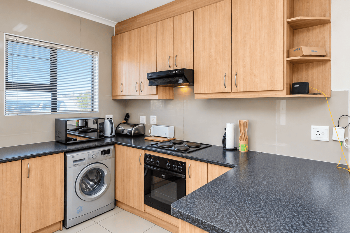 A functional kitchen featuring light wood-finish cabinetry, dark laminate countertops, a built-in oven with hob, and integrated laundry appliance space.