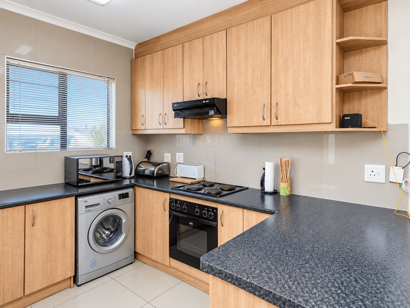 A functional kitchen featuring light wood-finish cabinetry, dark laminate countertops, a built-in oven with hob, and integrated laundry appliance space.