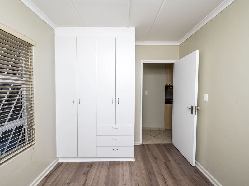 A clean, empty bedroom featuring modern laminate flooring, built-in white cupboards, and venetian blinds, offering a neutral canvas for personalization.