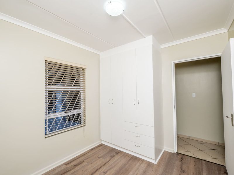 A clean, empty bedroom featuring modern laminate flooring, a built-in white wardrobe, and a window fitted with venetian blinds.