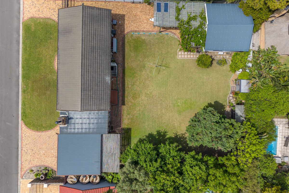 Aerial view of the property showcasing the main dwelling, expansive lawn, mature garden landscaping, paved driveway, and roof-mounted solar panels.