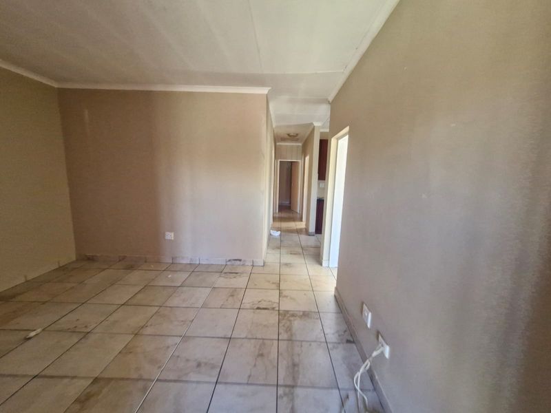 An empty living area featuring ceramic floor tiles and neutral wall paint, leading into a hallway with access to other rooms.