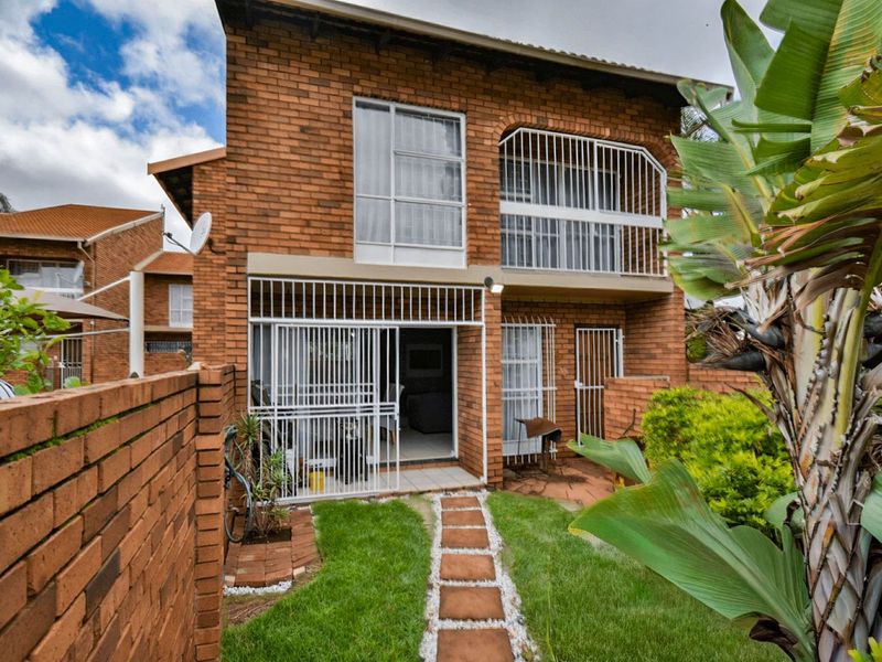 Rear view of a double-story face brick townhouse featuring a private garden, paved walkway, ground floor patio, and an upper-level balcony with security gates.