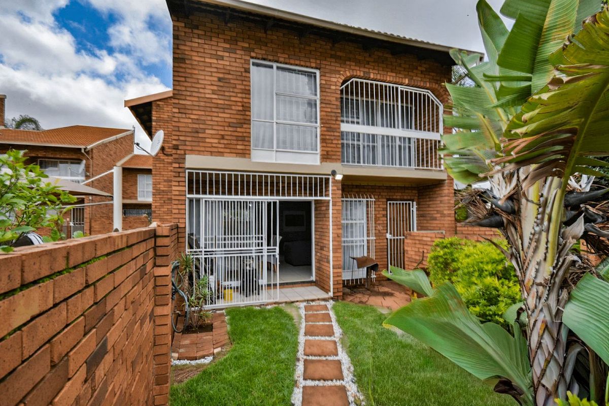 Rear view of a double-story face brick townhouse featuring a private garden, paved walkway, ground floor patio, and an upper-level balcony with security gates.