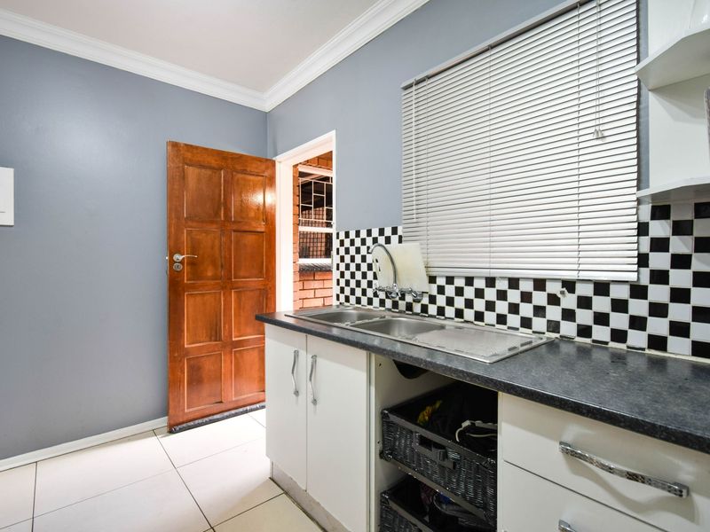 A functional scullery area featuring a double stainless steel sink, checkered tile backsplash, built-in cabinetry, and tiled flooring with direct access to an exterior space.