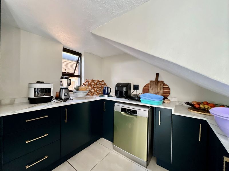 A compact, modern kitchen featuring dark cabinetry, stone countertops, and an integrated dishwasher, situated under a sloped ceiling with a window.