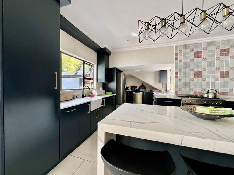 Modern kitchen featuring dark cabinetry, a large quartz-topped island, farmhouse sink, gas range, and decorative tiled backsplash with contemporary pendant lighting.