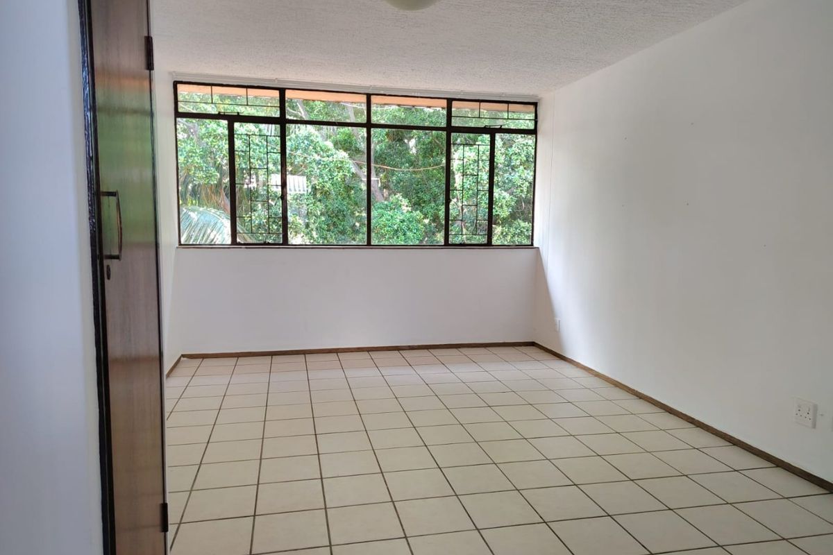A bright, empty bedroom featuring tiled flooring, a built-in cupboard, and large windows offering a pleasant leafy outlook.