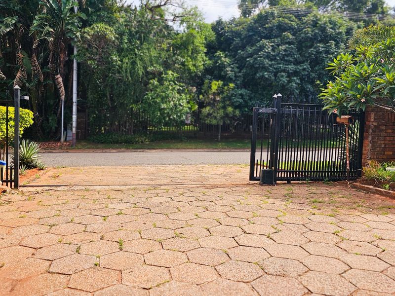 A view of the property entrance featuring a paved driveway, metal security gate, and established garden surroundings.