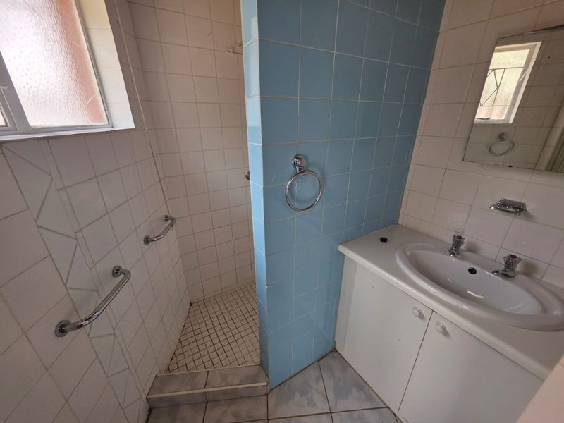 A dated bathroom featuring a shower cubicle, vanity cabinet with integrated basin, and wall-mounted grab rails, requiring cosmetic updates to modernize the space.