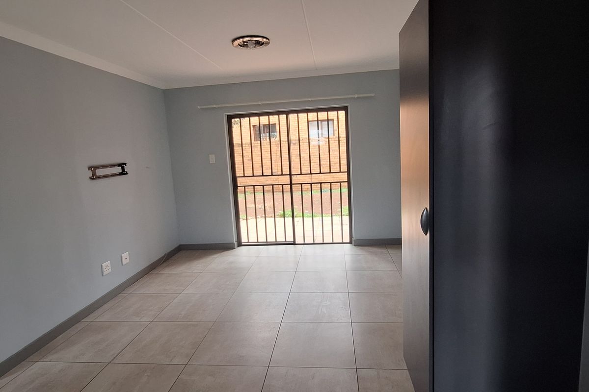 An empty living room featuring neutral tiled flooring, a sliding glass door with security gate, and a wall-mounted TV bracket.