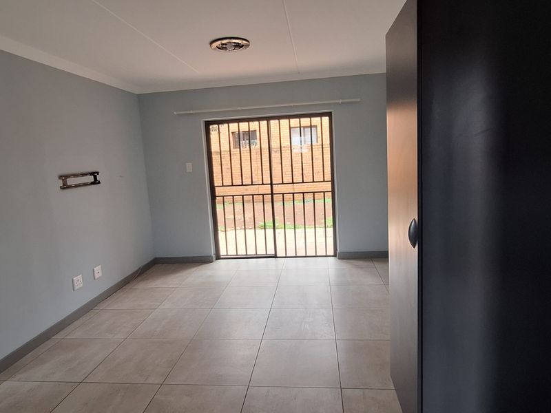 An empty living room featuring neutral tiled flooring, a sliding glass door with security gate, and a wall-mounted TV bracket.