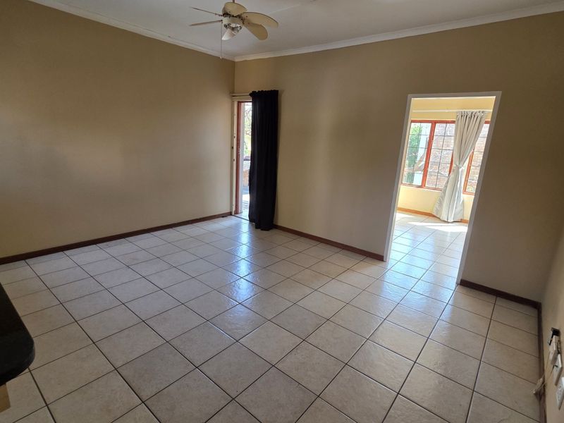 An empty, neutrally painted living area featuring tiled flooring, a ceiling fan, and an open doorway leading to an adjacent space.