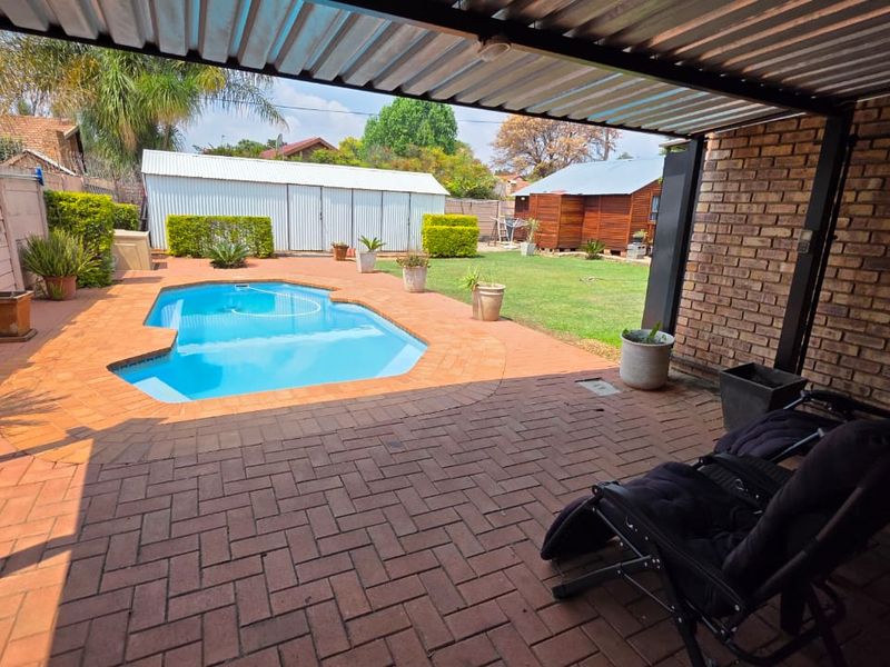Outdoor entertainment area featuring a covered patio with brick paving, overlooking a swimming pool and a well-maintained garden.