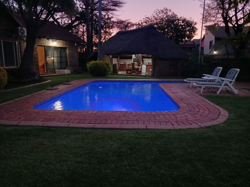 A well-maintained backyard featuring a swimming pool with brick coping, surrounded by a lawn and a traditional thatched lapa entertainment area.