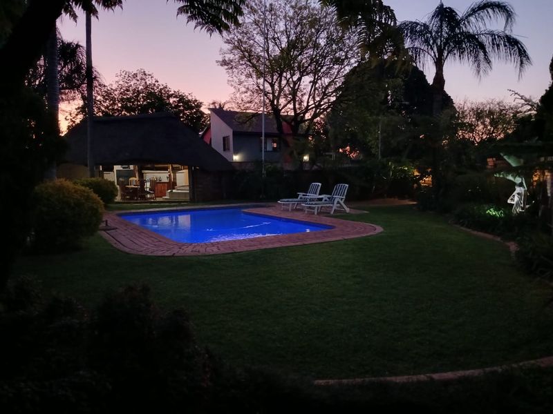 A well-maintained backyard featuring a swimming pool with brick coping and an adjacent thatched lapa, set within a landscaped garden at dusk.