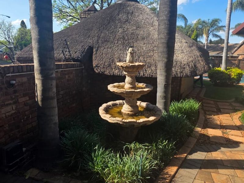 A landscaped garden area featuring a tiered stone fountain, paved pathways, and a traditional thatch-roofed structure in the background.