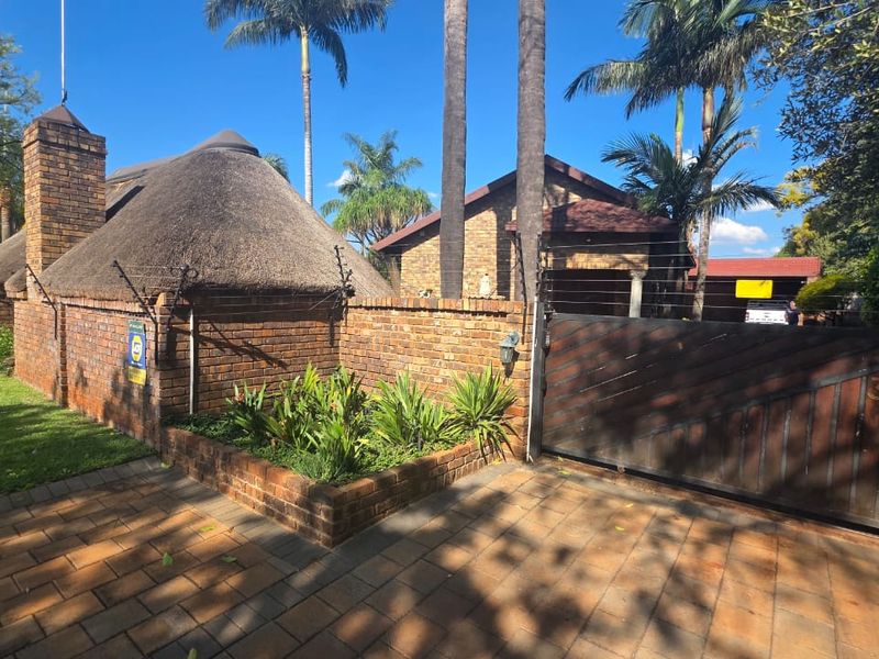 Front exterior view featuring a brick boundary wall, thatched roof structure, paved driveway, and secure sliding gate with electric fencing.
