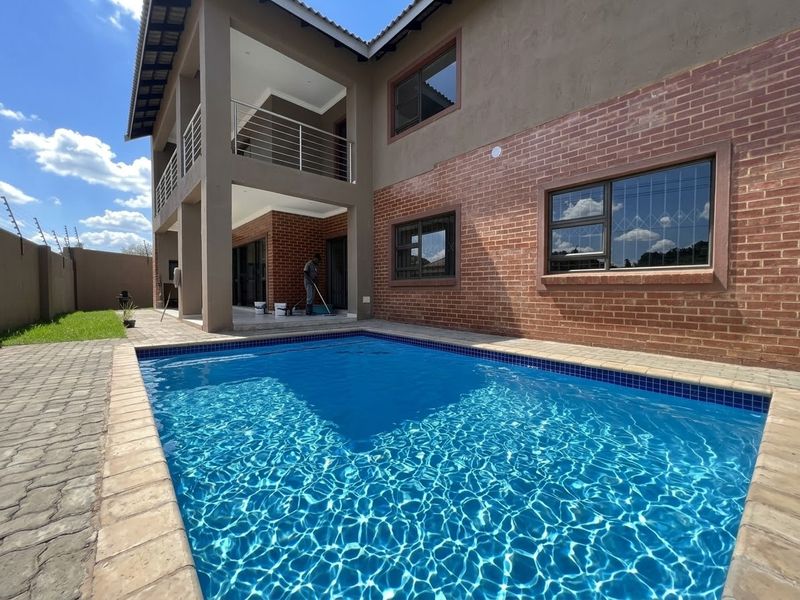 A well-maintained swimming pool area featuring a paved surround, brick exterior facade, and a covered patio with an upper-level balcony.