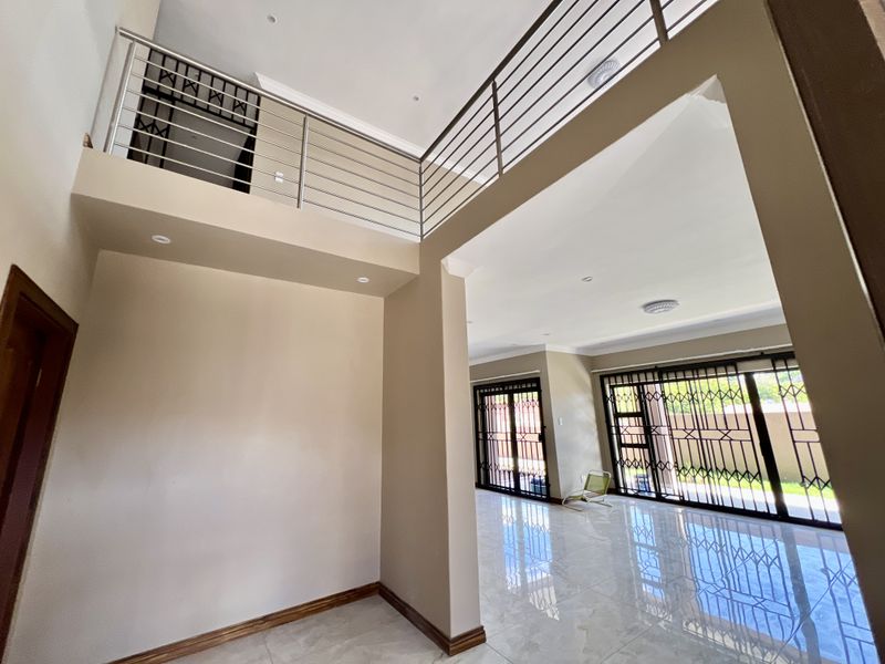 Spacious double-volume living area featuring tiled floors, recessed lighting, and a mezzanine level with a modern stainless steel balustrade, leading to outdoor access.