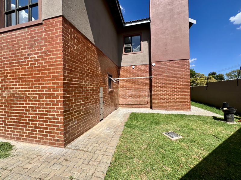 A well-maintained rear exterior view featuring exposed brickwork, a paved walkway, a neat lawn, and secure perimeter walling with electric fencing.