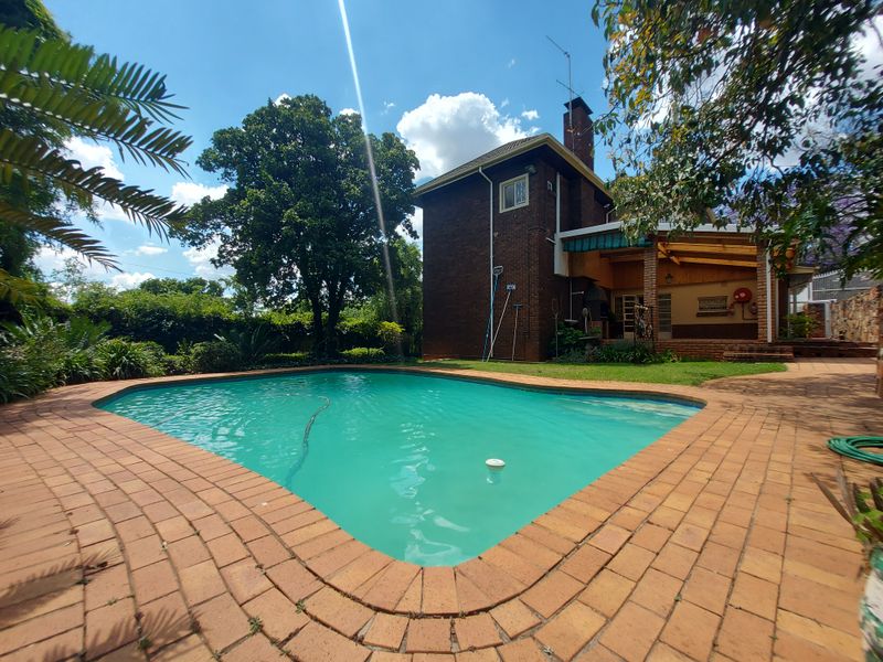 A well-maintained outdoor area featuring a swimming pool, brick paving, and a covered patio with a built-in braai, set within a landscaped garden.
