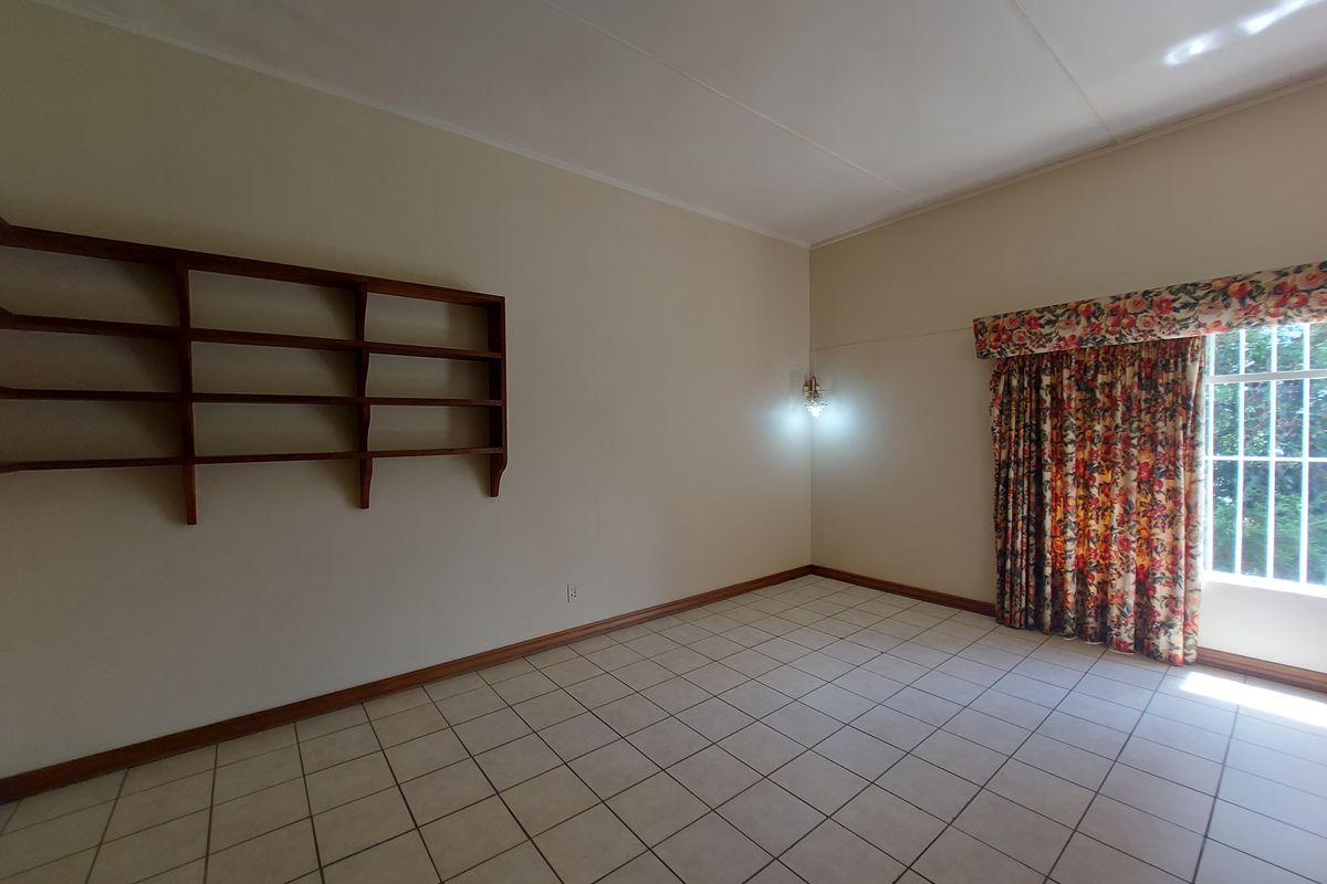 A spacious, empty bedroom featuring neutral tiled flooring, built-in wooden wall shelving, and a window with security bars.