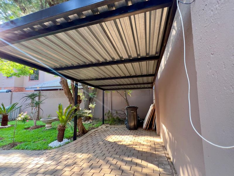 A covered outdoor patio area featuring a corrugated metal roof, steel support structure, and paved flooring, adjacent to a garden space.