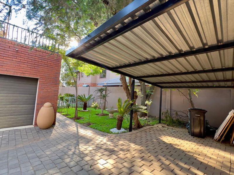A covered outdoor carport area featuring a corrugated metal roof, paved flooring, and adjacent garden space with a brick garage structure.
