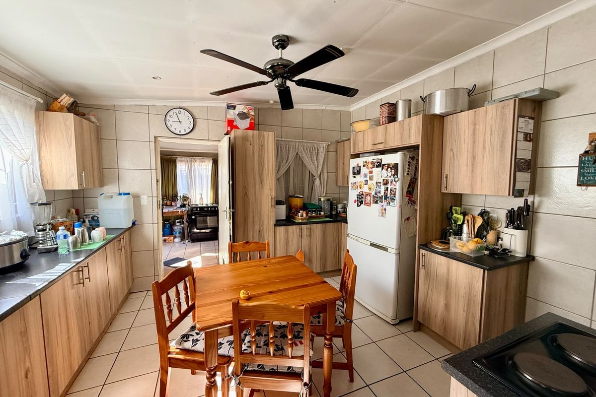 A functional kitchen featuring wood-look cabinetry, tiled walls and flooring, and a central ceiling fan, with an adjacent scullery area visible.