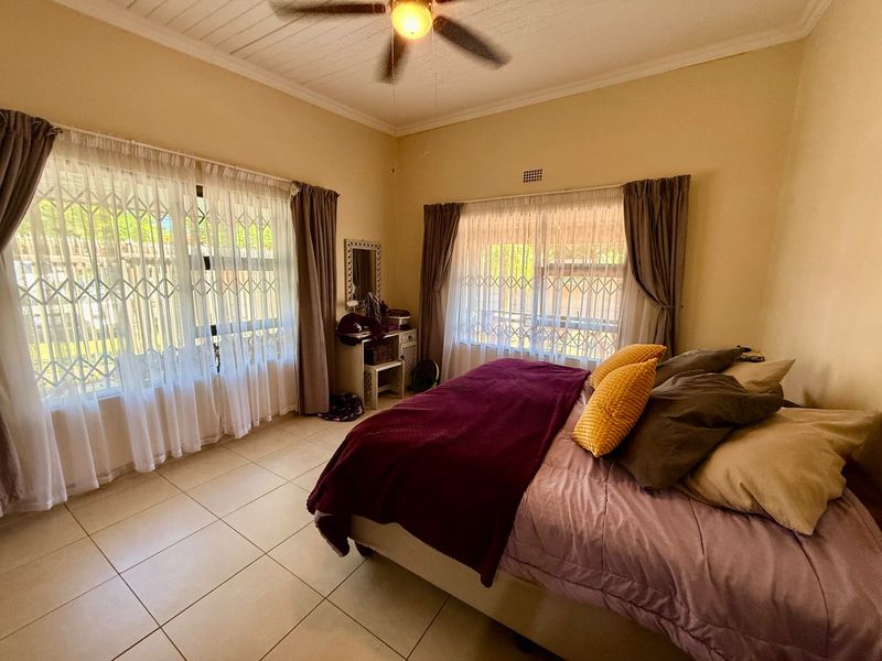 A well-lit bedroom featuring tiled flooring, a ceiling fan, and large windows fitted with security gates, offering a comfortable and functional living space.