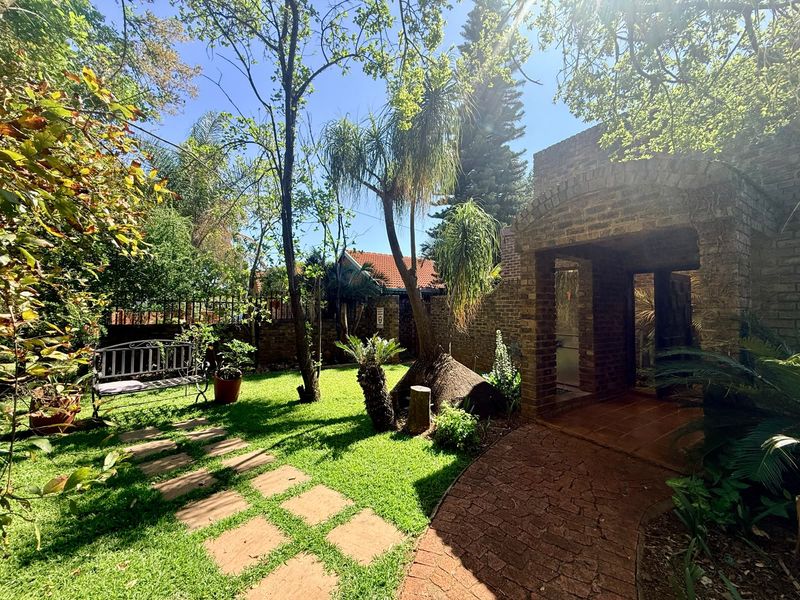 A lush, well-established garden featuring a brick-paved pathway leading to an arched brick entryway, surrounded by mature trees and greenery.