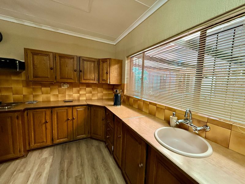A functional kitchen featuring traditional wooden cabinetry, laminate countertops, a tiled backsplash, and a large window with venetian blinds.