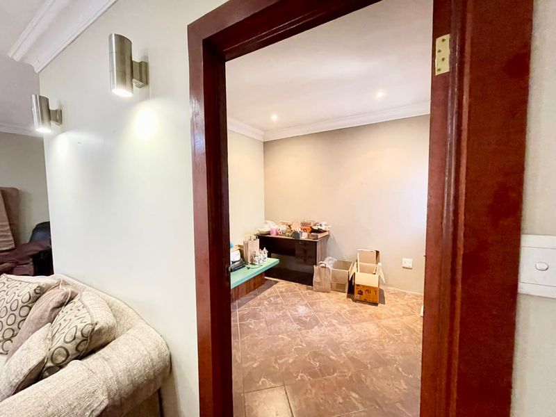 A functional office space featuring tiled flooring, recessed downlighting, and a built-in desk, viewed through a wooden door frame from an adjacent living area.