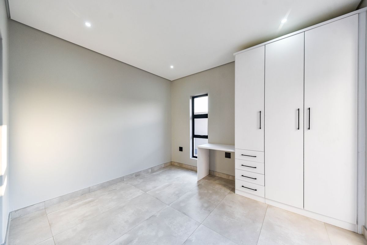A modern, empty bedroom featuring neutral tiled flooring, recessed lighting, and a sleek built-in wardrobe with an integrated desk area.