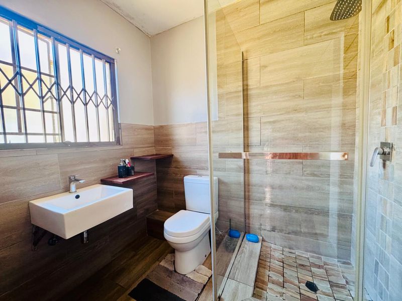 Modern bathroom featuring a wall-mounted basin, toilet, and a spacious glass-enclosed shower with wood-look wall tiling and security bars on the window.