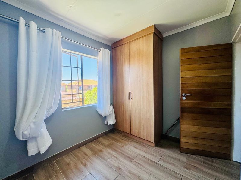A bright, empty bedroom featuring timber-look tiled flooring, a built-in wooden wardrobe, and a window with security bars.