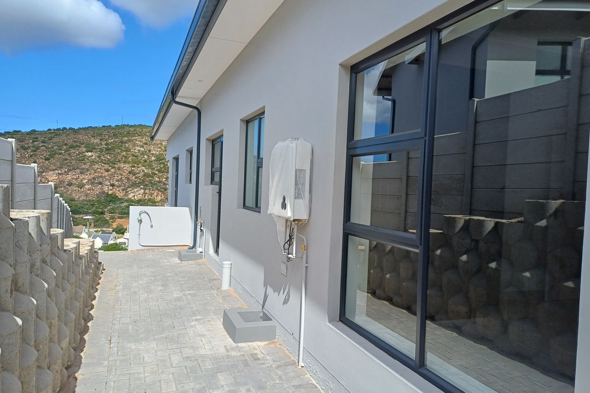 A side exterior view of the property featuring paved walkways, modern aluminium window frames, and a wall-mounted gas geyser unit.