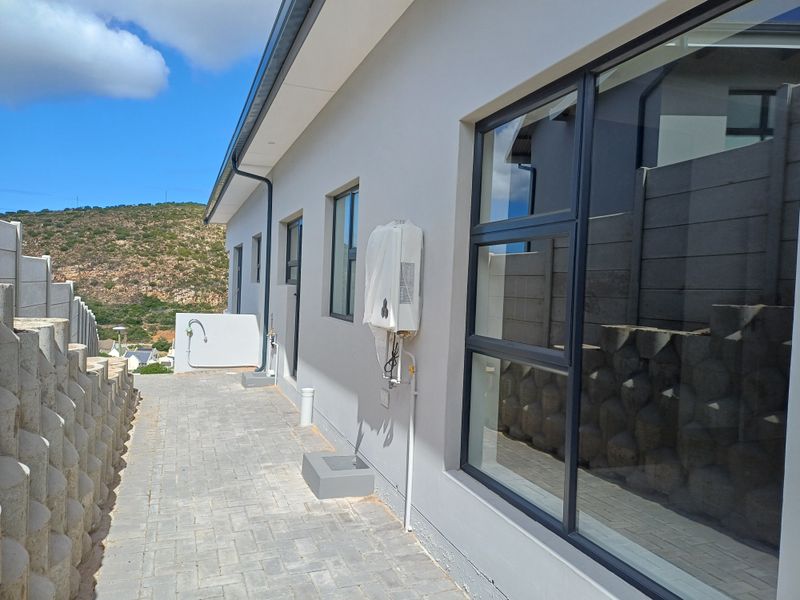 A side exterior view of the property featuring paved walkways, modern aluminium window frames, and a wall-mounted gas geyser unit.