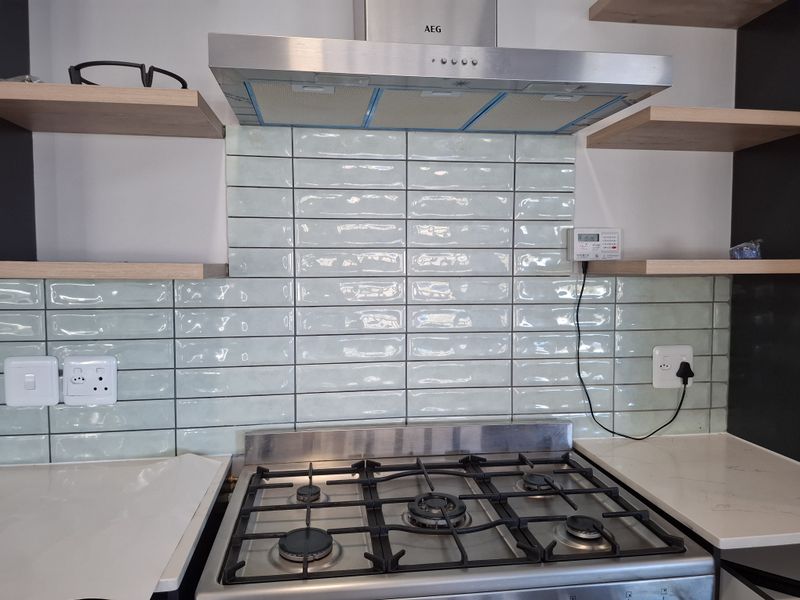 Modern kitchen area featuring a gas hob, stainless steel extractor fan, and stylish light green subway tile backsplash with floating wooden shelving.