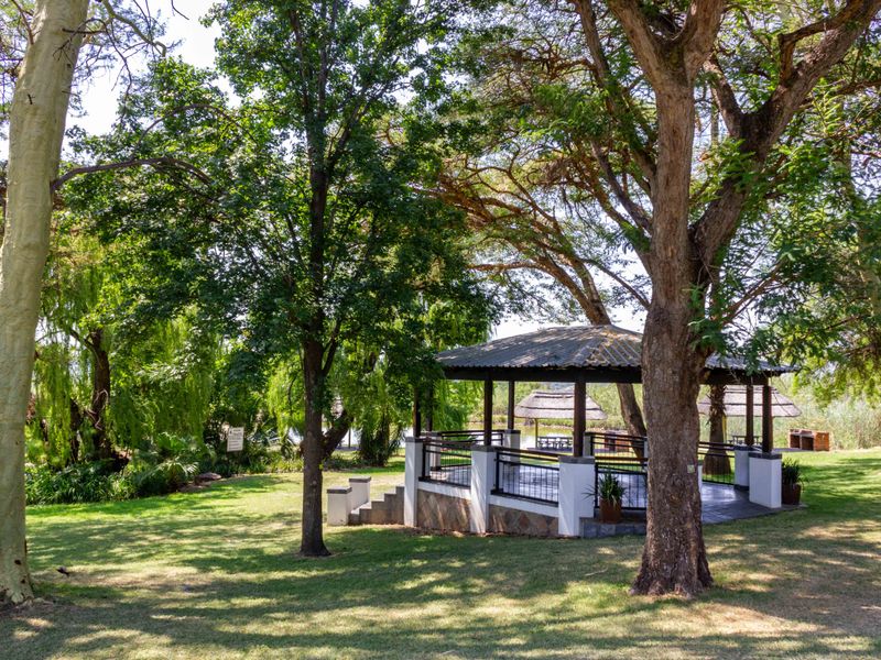 A well-maintained outdoor gazebo featuring tiled flooring, metal railings, and stone cladding, set within a lush, tree-filled garden environment.