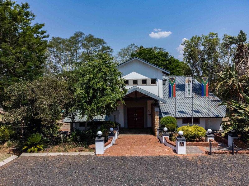 Front elevation of a lodge building featuring a metal roof, brick paved entrance path, stone accents, and established garden surroundings.