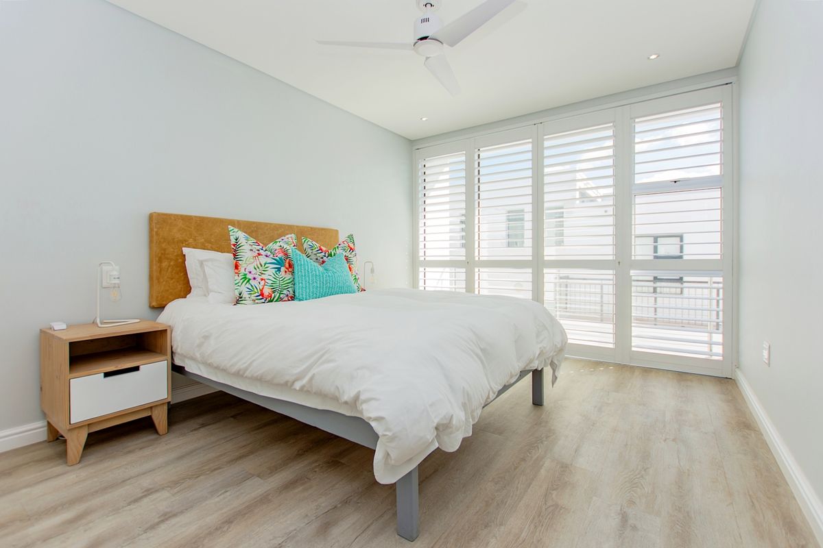 A bright and modern bedroom featuring light wood-look laminate flooring, a ceiling fan, and white plantation shutters allowing for natural light.