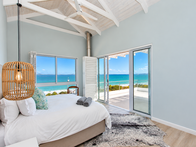 Bright bedroom featuring vaulted ceilings with exposed beams, laminate flooring, and large sliding glass doors opening to a balcony with panoramic ocean views.