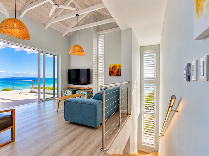 Bright living area featuring vaulted ceilings with exposed beams, modern laminate flooring, and large sliding doors opening to a scenic balcony with ocean views.