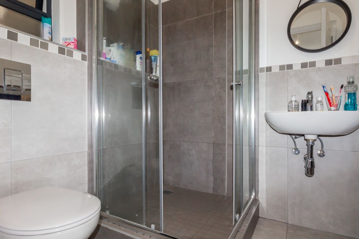 A compact modern bathroom featuring a glass shower enclosure, wall-mounted basin, and wall-mounted toilet with neutral grey tiling.