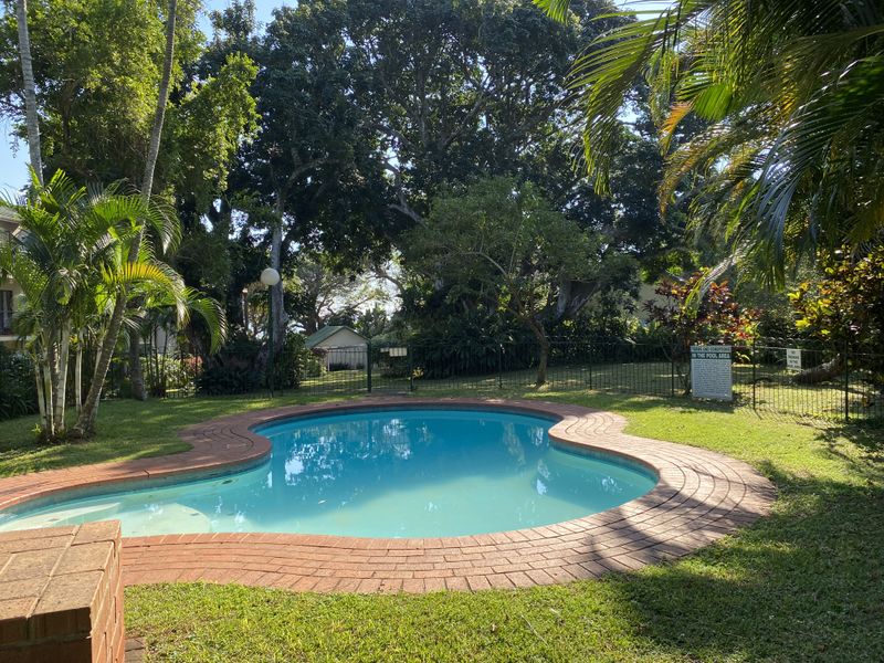 A well-maintained communal swimming pool area featuring brick coping, surrounded by lush established trees and landscaped garden space.