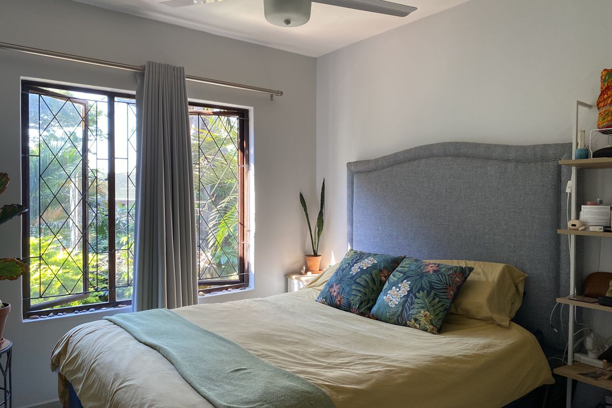 A well-lit bedroom featuring a ceiling fan and large windows with security bars, offering a view of the surrounding greenery.