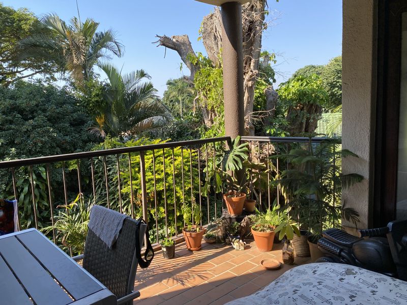 A covered balcony featuring tiled flooring and a metal balustrade, overlooking a lush, mature garden setting.