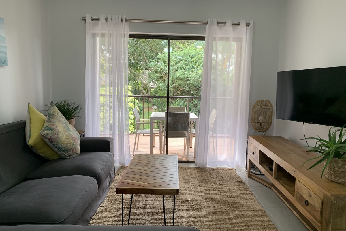 A bright living room featuring tiled floors and a large sliding glass door that opens onto a private balcony with garden views.